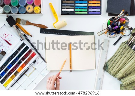 Artist drawing graphic sketch at sketchbook. Workplace, workspace. Top view photo of artistic tools lying on work-table: gouache, crayons palette and paintbrush collection.