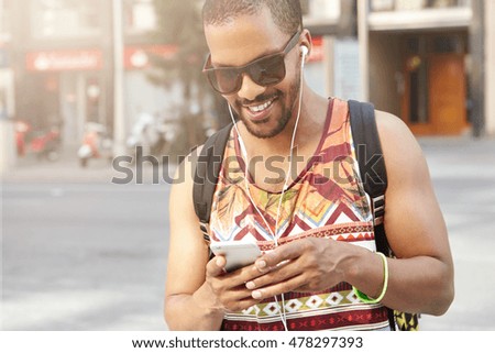 Muscular man in shades and stylish clothes, typing messages using wireless Internet connection on cell phone as walking down street, listening to new album of his favourite artist with headphones