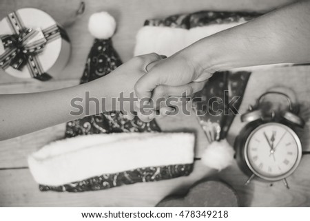 Black and white top view picture of male shaking female hand above table with gift box and Santa Claus hat. Closeup of holiday planning with alarm clock on wooden plank background.