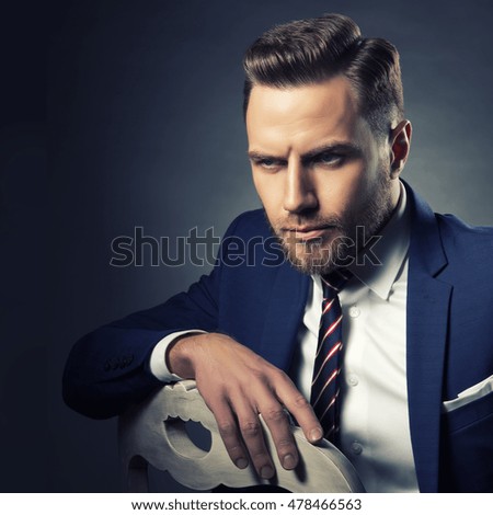 Young handsome bearded caucasian man with blue eyes sitting on chair. Perfect skin and hairstyle. Wearing blue suit. Studio portrait on gradient black to grey background. Toned
