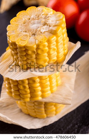 Vertical photo of three pieces of yellow sweet corn. Food is placed on a stack with leaves between. Corn is on black stale stone and wooden board with tomatoes and bread.