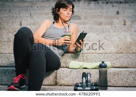 Woman drinking juice vegetable green detox cleanse smoothie after fitness running workout listening music using her phone with bottle of water and towel. Healthy lifestyle concept.