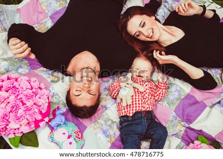 Smiling parents in black t-shirts and little boy in plaid shirt lie on the pink blanket