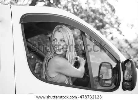 Black and white picture of beautiful blond woman driving car. Girl looking from car window at camera on blurred countryside background.