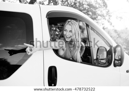 Black and white picture of pretty girl in car excited and looking from car window. Young woman smiling and pointing at something on road on summer countryside background.