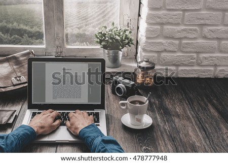 Hipster man working at home on a desk in front of a window, he is typing on a laptop and having a tea, point of view shot