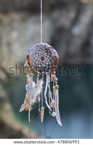 Handmade pink native american dream catcher on background of rocks and lake. Tribal elements, feathers, shells, lace