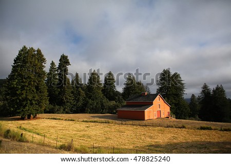 Big red barn in rolling farm land hills with forest trees and storm clouds at sunset