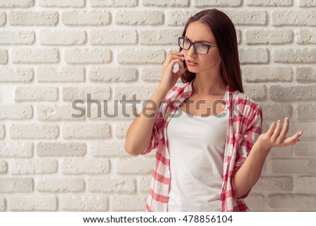 Attractive young girl in casual clothes and eyeglasses is talking on the mobile phone and gesturing, on white brick wall background