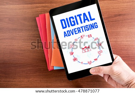 Top view of hand holding tablet with Digital advertising word with icon over color notebook on wooden table top,Digital business concept