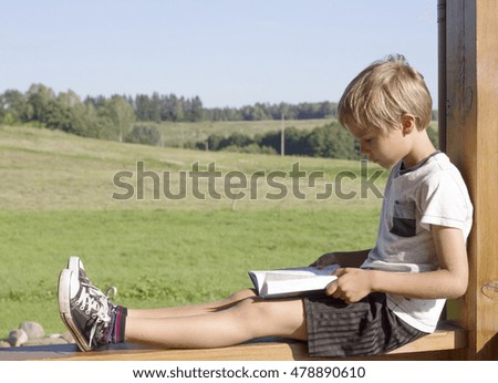 Little boy sitting and reading book at summer terrace. Casual clothes. Nature background. People, education, knowledge concept