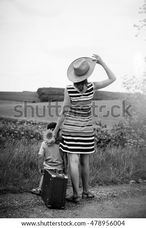 Black and white picture of woman and child on roadside near summer field. Kid sitting on suitcase and female holding sunflowers on sunset countryside background.