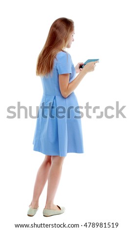 back view of standing young beautiful woman using a mobile phone or tablet computer. Isolated over white background. Skinny girl in blue dress playing the game on the tablet.
