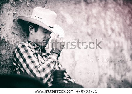 A cowboy sitting on a fence and leaning against a wall. Black and white, vintage photo with some scratches. Place for your text.