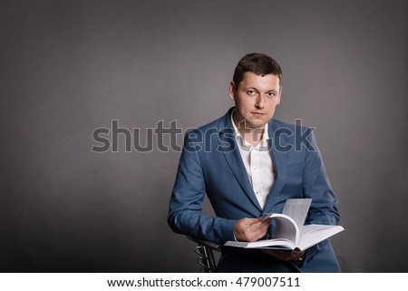 Portrait of extravagant and charming young man in the studio dark background in the official style of  blue suit is reading a book and holding in hands