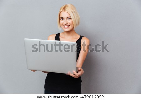 Smiling attrative young woman standing and using laptop over gray background