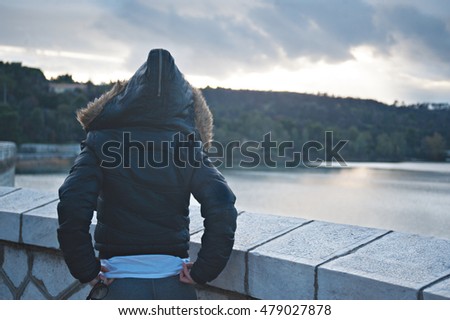 woman in back wearing the hood of here jacket up in a bridge and looking the river before the night is come