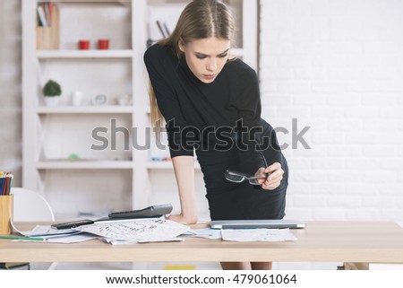 Pretty female designer leaning on desktop and drawing interior project. Shelves with various items in the background 