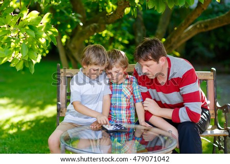 Two funny handsome kid boys and young father playing together checkers game. Sons, siblings children and dad spending leisure together. Family having fun in summer garden outside.