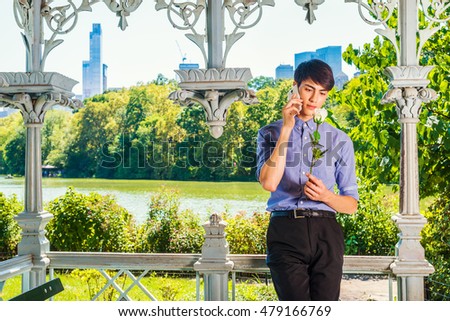 Love starts with Nature. 20 years old young Asian American college student stands inside pavilion by lake at New York Central Park in summer, holding white rose, talking on cell phone. I miss you.

