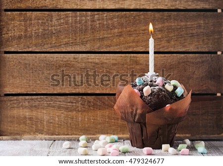 Delicious birthday chocolate muffin with colorful marshmallows and a single candle on a wooden background, selective focus. Toned image with copy space