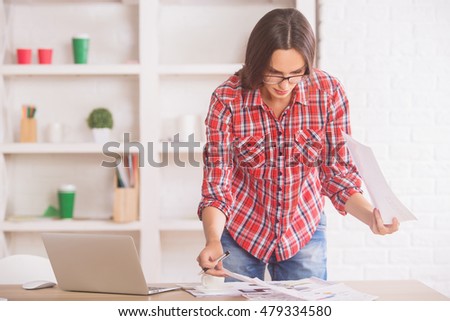 Beautiful young business woman standing at office desk with coffee cup, laptop, other items and doing paperwork