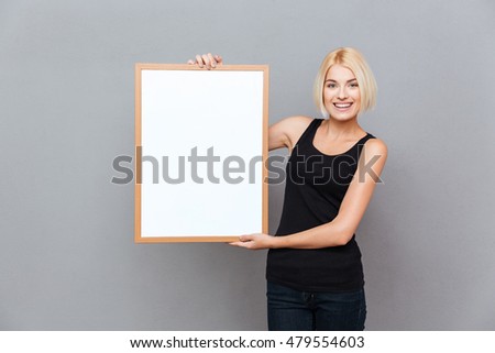 Happy pretty young woman holding blank white board over gray background