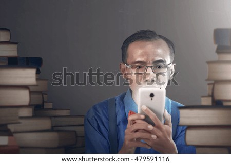 Nerdy man with serious expression holding smartphone, in empty room with stacking books background