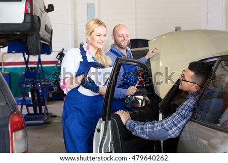 Man has brought his broken car to a repairshop. Beautiful female mechanic is writing up the symptoms while her male colleague is opening a hood