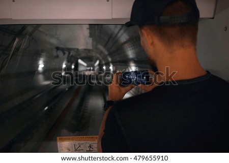 Young man in baseball hat and navy t-shirt taking a photo of the subway tunnel from the last car on his smartphone