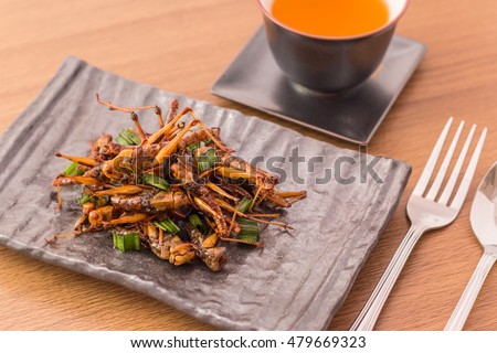 Set of Grasshopper edible insect crispy mixed with pandan, Thai pepper powder,  in a brown plate with fork and spoon, tea on wooden table background, Selective focus, Horizontal image