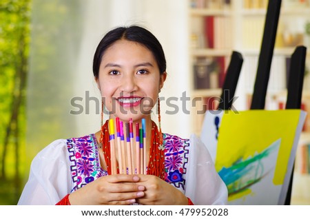 Young brunette woman wearing traditional andean clothing, holding up selection of color pencils and smiling happily, canvas behind, inside studio, garden window background