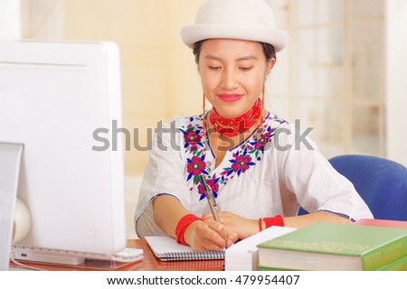 Young pretty girl wearing white shirt with colorful flower decorations and fashionable hat, sitting by desk working writing on paper smiling, stack of books, bright background