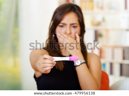 Pretty young brunette woman holding pregnancy home test, shocked facial expression covering mouth with hand, bookshelves interior, garden window background