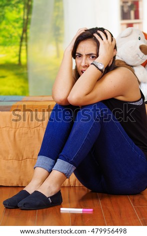 Pretty young brunette woman sitting on floor with pregnancy home test lying in front, looking shocked desperate, garden window background