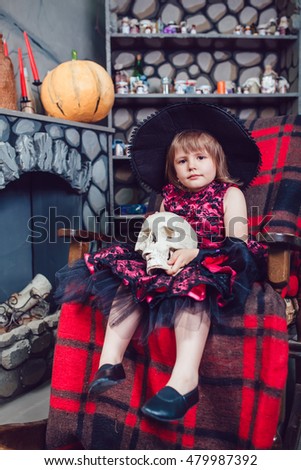 Girl in a witch costume sitting in a chair with a skull in his h0and in in halloween decorations