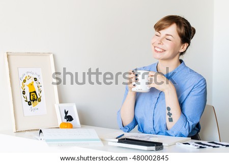 Beautiful young woman graphic designer / illustrator sitting at table with a cup in her hands, smiling widely, eyes closed