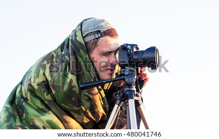 Closeup of a camouflaged paparazzi photographer  taking picture with a camera on a tripod
