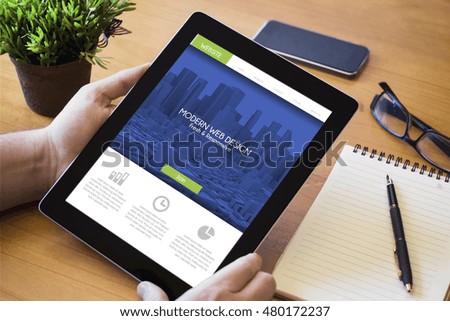 hands of a man holding a fresh design website on tablet device over a wooden workspace table. All screen graphics are made up.