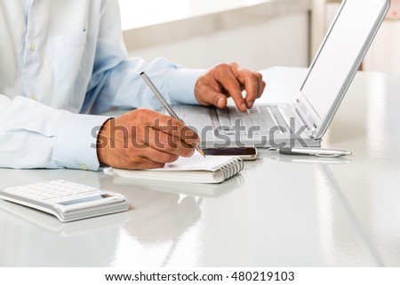 An Unrecognizable man is working by using a laptop computer on white desktop. Hands typing on a keyboard and writing on a blank sheet of notebook