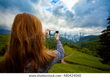 Young woman is standing on the hill and taking photo of amazing beautiful landscape with mountain, forest and cloudly sky