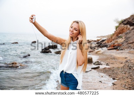 Young smiling beautiful woman listening music and making selfie while standing on the rocky beach