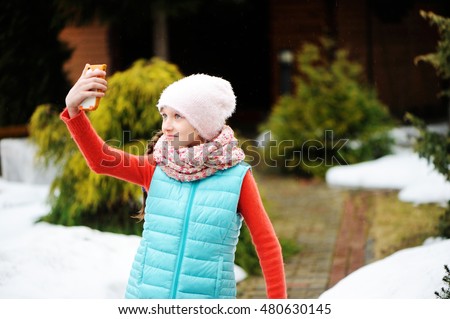 Adorable pre-teen tweenie kid girl in winter clothes making self portrait with funny face on her smartphone