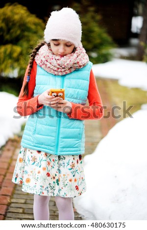 Adorable pre-teen tweenie kid girl in colorful winter clothes, hat and scarf with her smartphone in the winter park