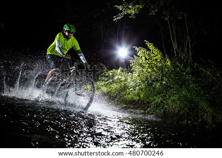 Mountain biker speeding through forest stream. Water splash in freeze motion. Night ride.