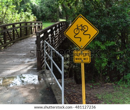 Yellow sign showing warning of slippery when wet alert caution next to wooden bridge over little creek in bike and hike trail of city park in Houston, Texas, US. Safety workout in urban park concept.