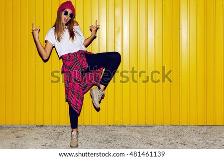 Young stylish hipster  girl having fun   and doing grimaces  near bright yellow urban wall at night with flash. Wearing pink hat, warm hipster boots.  Royalty-Free Stock Photo #481461139