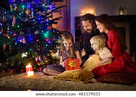 Young happy family of four unwrapping Christmas gifts by a fireplace in a cozy dark living room on Christmas eve