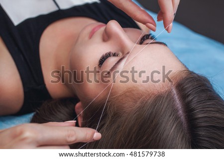 Master corrects makeup, gives shape and thread plucks eyebrows in a beauty salon. Professional care for face. Royalty-Free Stock Photo #481579738