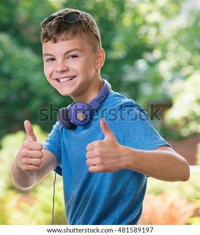 Portrait of a teen boy 12-14 year old showing thumb up gesture. Happy child having fun in park. Student with headphones and sunglasses on green background.